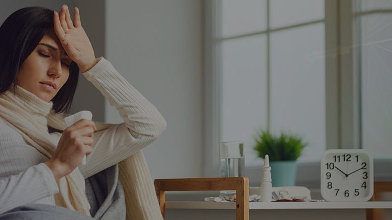 Woman checking temperature, sitting with medicine and clock on table.